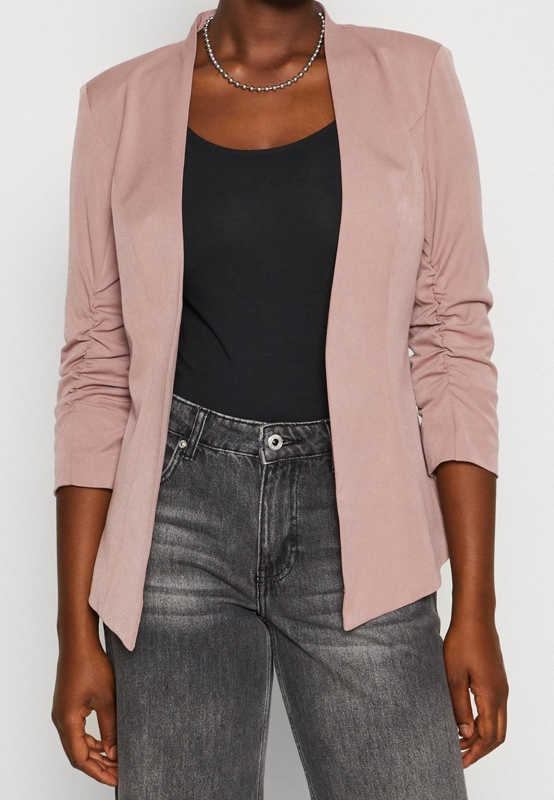 Femme portant un blazer rose poussiéreux à manches froncées sur un haut noir et un jean noir délavé taille haute, accessoirisée d'un collier de perles.