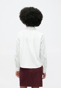 Person with curly hair wearing a white long-sleeve blouse with decorative pearls on the collar and a burgundy skirt, facing away.