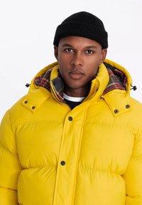 Gul puffer-jakke med aftageligt ternet for, sorte knapper og hætte. Stoffet ser skinnende ud med quiltet tekstur. Sort strikket hue bæres.