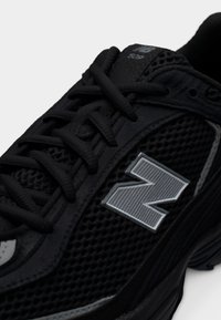 Μαύρο αθλητικό παπούτσι με επάνω μέρος από διχτυωτό υλικό, διαθέτει ασημένιο λογότυπο 'N', μαύρα κορδόνια και υφή στην εξωτερική σόλα για καλύτερη πρόσφυση και υποστήριξη.