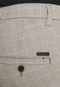 Beige mönstrade byxor i en texturerad tyg, med en svart knapp och en liten märkesetikett. Rutig design med varierande rutor.