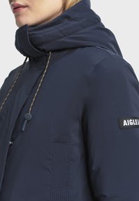 Navyblauwe geïsoleerde jas met een hoge kraag, gestikte details, zijzakken en een logo-opdruk op de mouw. Trekkoord bij de hals.