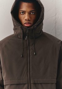 Bruine capuchonjas met ritssluiting, trekkoord en een boxy silhouet. Soepele textuur met naadtoevoegingen op mouwen en lijf.