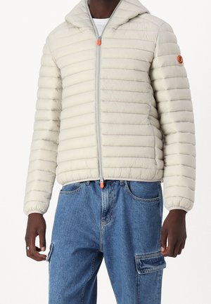 Mężczyzna w jasnobeżowej pikowanej kurtce z kapturem, wyposażonej w pomarańczowe suwaki, oraz w niebieskie dżinsowe spodnie cargo na białym tle.