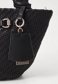 Czarna pleciona torba na ramię z skórzanymi akcentami, z metalowym uchwytem w kolorze złotym oraz prostokątną metką z wytłoczonym napisem "GUESS".