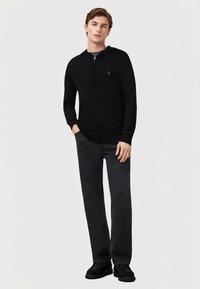 Crna dukserica s patentnim zatvaračem od mekanog materijala, s prednjim džepom i minimalističkim logom. Uparena s tamnosivim hlačama i crnim cipelama.