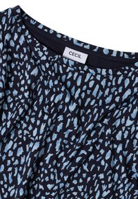 Marineblaue Stoff mit unregelmäßigen hellblauen Flecken, die ein Leopardenmuster bilden, und einem weißen Etikett mit der Aufschrift "CECIL" am Ausschnitt.