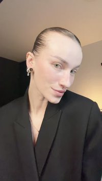 Črni blazer z globokim V-izrezom, urejeni lasje in svetla polt. Očiten obročasti in številčni uhani z detajli. Zelene oci.