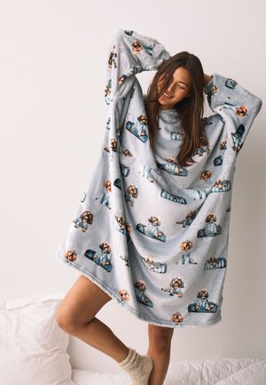 Smilende kvinde, der strækker sig i en stor lysegrå fleeceplaid med tegnefilmshund og skateboardprint, stående på sengen iført sokker.