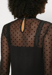 Schwarzes Kleid mit durchsichtigen Pünktchenärmeln, Spitzenakzenten an den Schultern und einem tropfenförmigen Schlüssellochverschluss hinten.