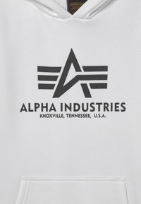 Bílá mikina s kapucí s výrazným černým logem se ''A'' a horizontálními pruhy. Text zní "ALPHA INDUSTRIES KNOXVILLE, TENNESSEE, U.S.A."