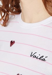 Camiseta blanca con rayas horizontales rosas, adornada con corazones de lentejuelas rojas y texto bordado en negro "Voilà" en el pecho.