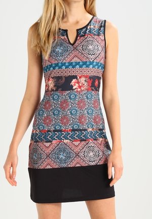 Mujer que lleva un vestido sin mangas con patrones mixtos de flores y geométricos en rojo, azul, negro y beige, y sección de falda negra sólida.