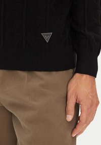 Schwarzer Strickpullover mit strukturiertem Muster, gerippten Bündchen und Saum. Auf der linken Seite befindet sich ein dreieckiges graues Logofähnchen. Beige Hose dargestellt.