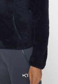 Pullover en polaire navy avec une texture douce, comportant une poche zippée sur le côté et un pantalon de sport gris contrastant avec un détail zippé.