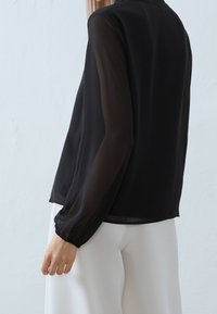 Blusa nera con maniche lunghe trasparenti, scollatura rotonda e polsini aderenti. Tessuto leggero con una consistenza liscia. Abbinata a pantaloni bianchi.