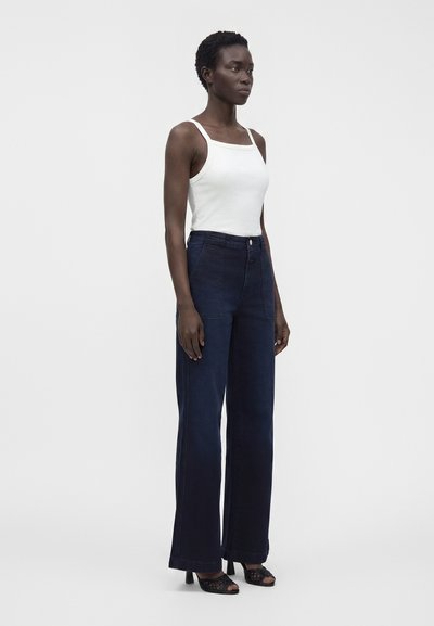 Femme debout, portant un haut blanc sans manches, un pantalon large taille haute bleu foncé et des chaussures noires à talons ouverts à l'avant, sur un fond uni.