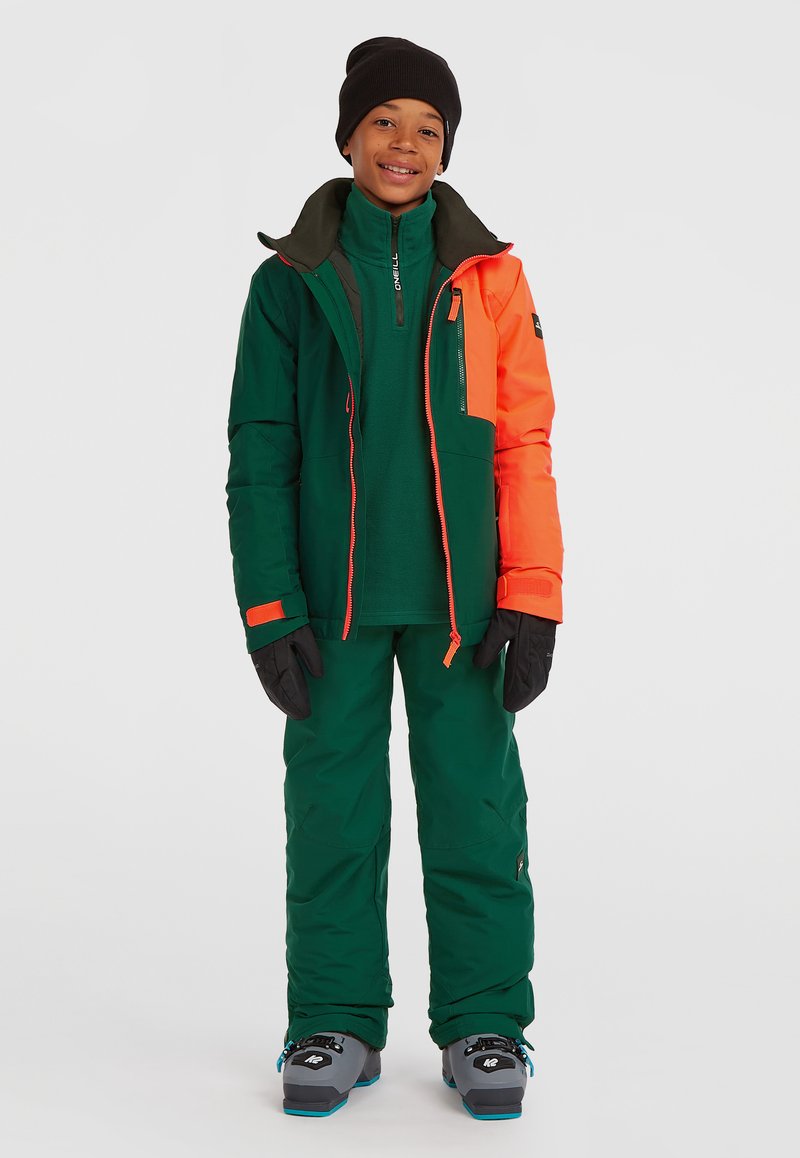 Groene en oranje geïsoleerde skijas met bijpassende broek, met een hoge kraag, ritszakken en zwarte handschoenen. Duurzaam, weerbestendig materiaal.