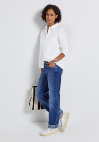 Femme aux cheveux bouclés se tient contre un mur blanc, portant une chemise blanche à boutons, un jean bleu, des baskets blanches, une montre en argent, et tenant un sac rayé.
