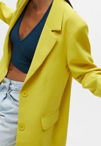 Personne portant un blazer oversize jaune vif sur un débardeur court vert sarcelle foncé et un jean taille haute bleu clair.