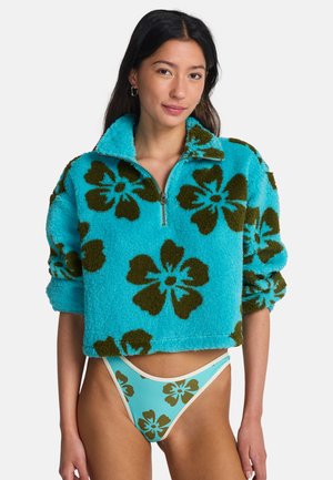 Kobieta ubrana w turkusowy polar z dużymi brązowymi kwiatowymi wzorami oraz dopasowane, wysokie majtki bikini.