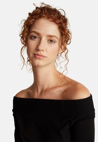 Černý svetr bez ramínek s žebrovaným výstřihem, s měkkou pletenou texturou. Modelka má kudrnaté, červené vlasy a přirozený make-up.