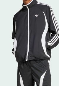 Svart sportjacka med vita ränder och detaljer, hög krage, dragkedja och elastiska muddar, kombinerad med matchande byxor med liknande design.