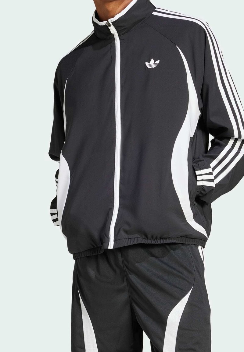 Svart sportjacka med vita ränder och detaljer, hög krage, dragkedja och elastiska muddar, kombinerad med matchande byxor med liknande design.