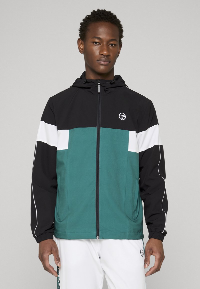 Noor mees, kes kannab musta, valget ja sinakasrohelist kapuutsiga lukuga jopet ning valgeid pükse, millel on rinna ja vasaku reie peal logo, seisab halli tausta ees.