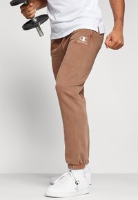 Brune bomulds sweatpants med elastiske manchetter, der har et hvidt Champion-logo på venstre lår, parret med hvide sportsko.