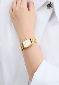 Goldfarbener Uhr mit weißem Zifferblatt, rechteckigem Gehäuse, römischen Ziffern und einem gewebten Metallarmband, getragen am Handgelenk über einem weißen Hemd.
