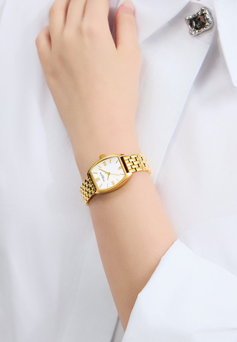 Goldfarbener Uhr mit weißem Zifferblatt, rechteckigem Gehäuse, römischen Ziffern und einem gewebten Metallarmband, getragen am Handgelenk über einem weißen Hemd.