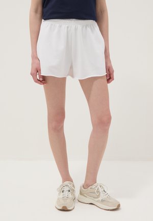 Personne portant un short blanc à taille élastique, un haut bleu marine et des chaussures de sport beiges, debout devant un fond clair uni.