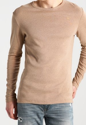 Homme portant une chemise beige ajustée à manches longues et un jean bleu déchiré, debout avec une main dans la poche sur un fond uni.