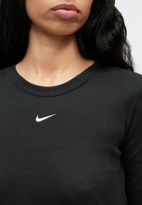 Μαύρο μπλουζάκι με μακριά μανίκια και ριμπ ύφασμα, με λευκό λογότυπο Nike στο στήθος. Η λαιμόκοψη είναι στρογγυλή και εφαρμοστή, με υφή στο ύφασμα.