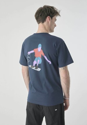 Man facing sideways wearing a navy shirt with a graphic of a purple-skinned figure skateboarding on a computer keyboard.