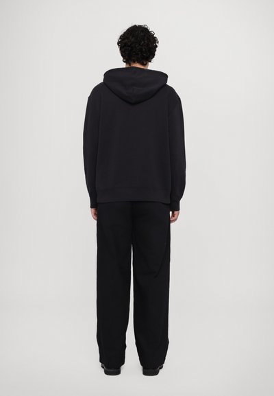 Haut à capuche noir surdimensionné, avec des épaules tombantes et des poignets côtelés, associé à un pantalon noir ample. Texture lisse.
