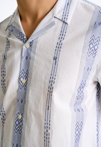 Camisa de manga corta en tejido blanco con patrones geométricos azules, cuello con muesca y cierre frontal con botones. Acabado texturizado.
