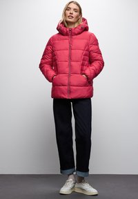 Růžová péřová bunda, prošívaný design s kapucí, přední zip a boční kapsy. V kombinaci s tmavými džíny a bílými teniskami.