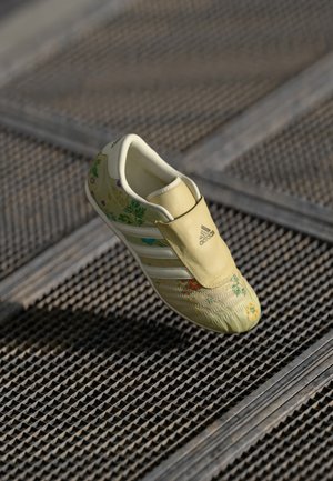 Zapatilla Adidas amarilla con patrones florales y rayas blancas sobre suelo de rejilla metálica.