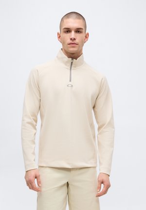 Junger Mann mit kurzen Haaren, der einen beigefarbenen Pullover mit kurzem Reißverschluss und passende Hosen vor einem schlichten hellen Hintergrund trägt.