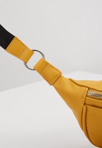 Marsupio in ecopelle gialla con cerniera metallica, cinturino nero regolabile e un anello circolare in argento. Texture liscia.