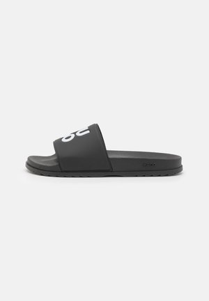Black slide sandals with a wide strap featuring a white logo. Made of durable material, they have a textured outsole for grip.
