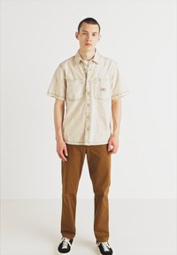 Short-sleeve beige shirt with a button-down front and two chest pockets, paired with brown pants and black sneakers on a plain background.