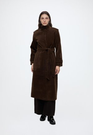 Femme debout portant un long manteau en velours marron foncé à col montant, taille ceinturée, pantalon large noir et mocassins noirs.