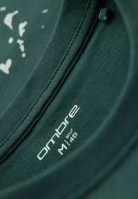 Mørkegrøn stof t-shirt krave med hvidt mærke, der viser "ombre" og størrelse "M" (EUR 48). Glat tekstur med rund halsudskæring.