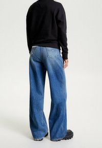 Jeans in denim blu a gamba larga con una texture sbiadita, tasche posteriori cucite e orli grezzi, abbinati a una felpa nera e scarpe con suola in gomma.