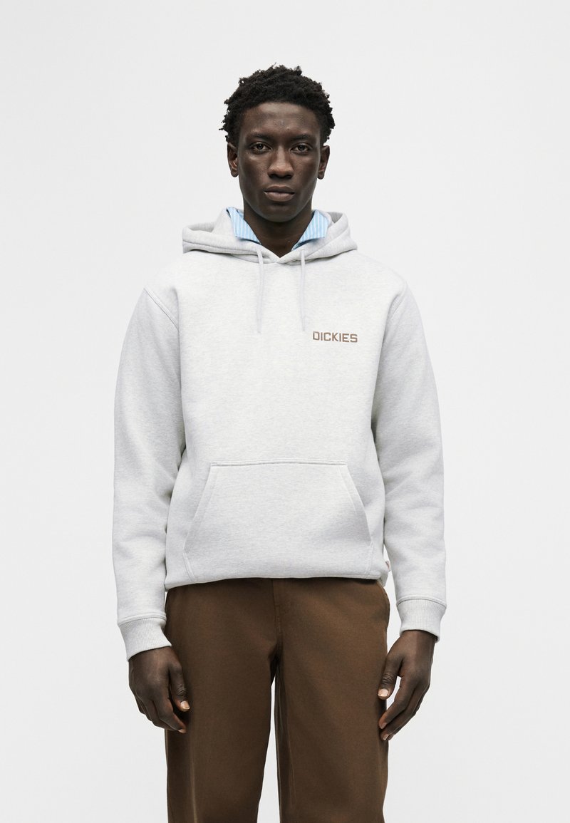 Giovane uomo con felpa con cappuccio grigio chiaro con logo "Dickies", pantaloni marroni e camicia blu con colletto sotto, in piedi davanti a uno sfondo neutro.