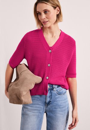 Kvinde iført en knaldrød strikket cardigan med korte ærmer og sølvknapper, lyseblå jeans og med en lysebrun ruskinds taske under armen.