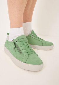 Pies con zapatillas de ante verde claro y suelas blancas, con cremalleras laterales, combinadas con calcetines blancos hasta el tobillo sobre un fondo neutro.